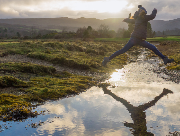 Girl in jacket and woolly hat jumping over stream