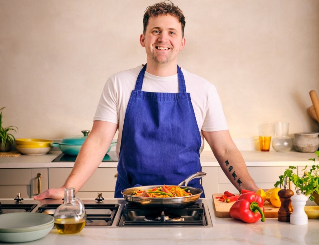 Jon Watts standing in a kitchen over a stovetop with a pan wearing a blue apron over a white t-shirt.