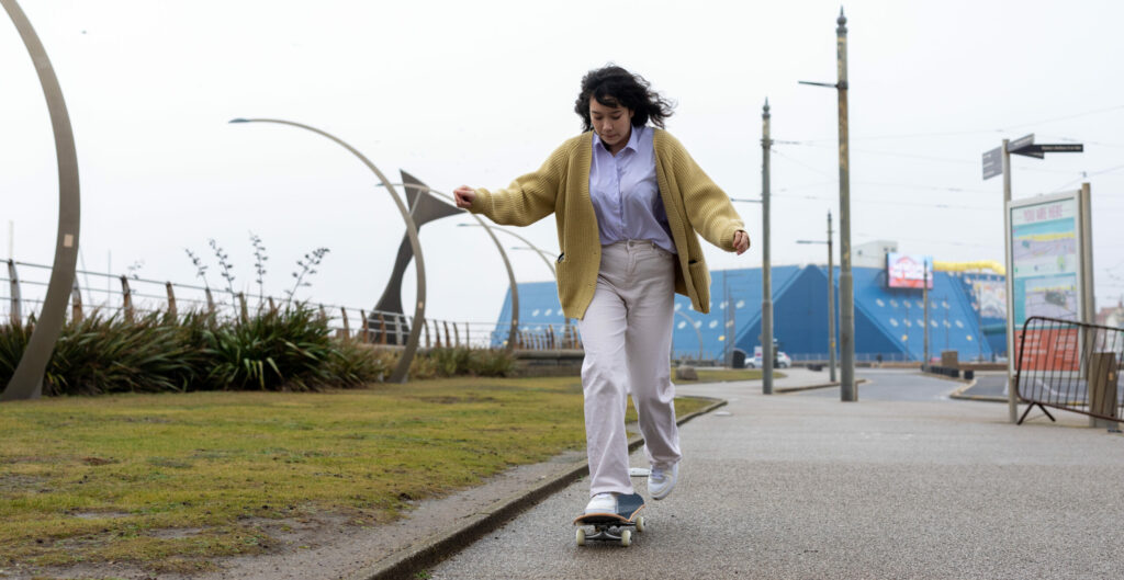A young person wearing a yellow cardigan and white trousers skateboarding down the street.