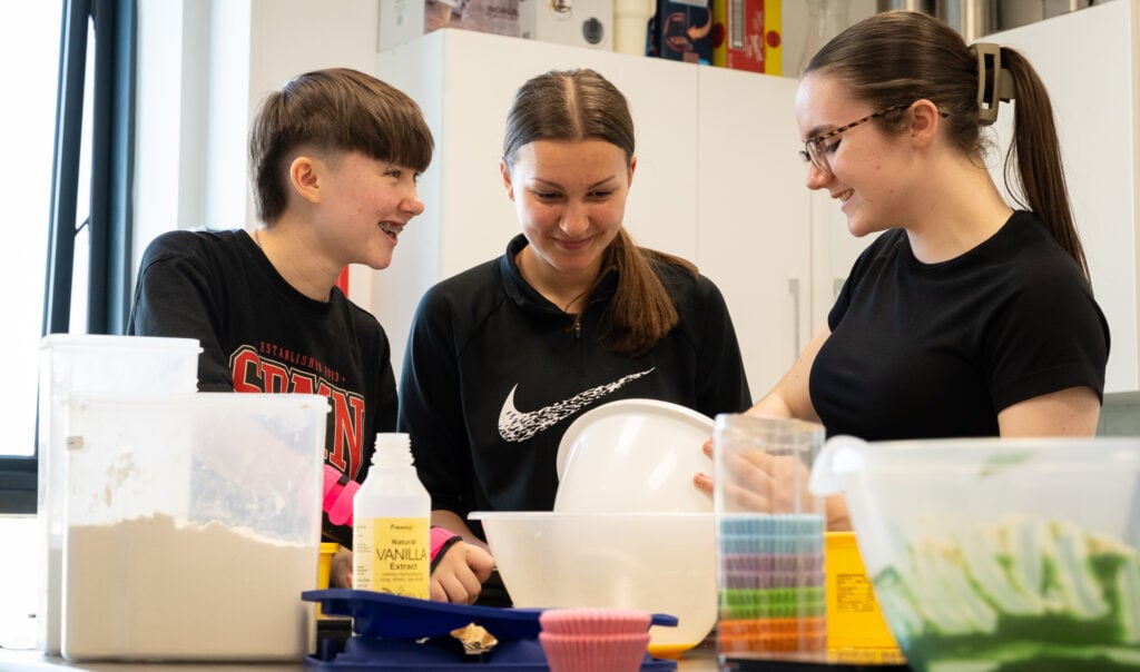 Three people looking at each other smiling standing behind ingredients and equipment for baking.