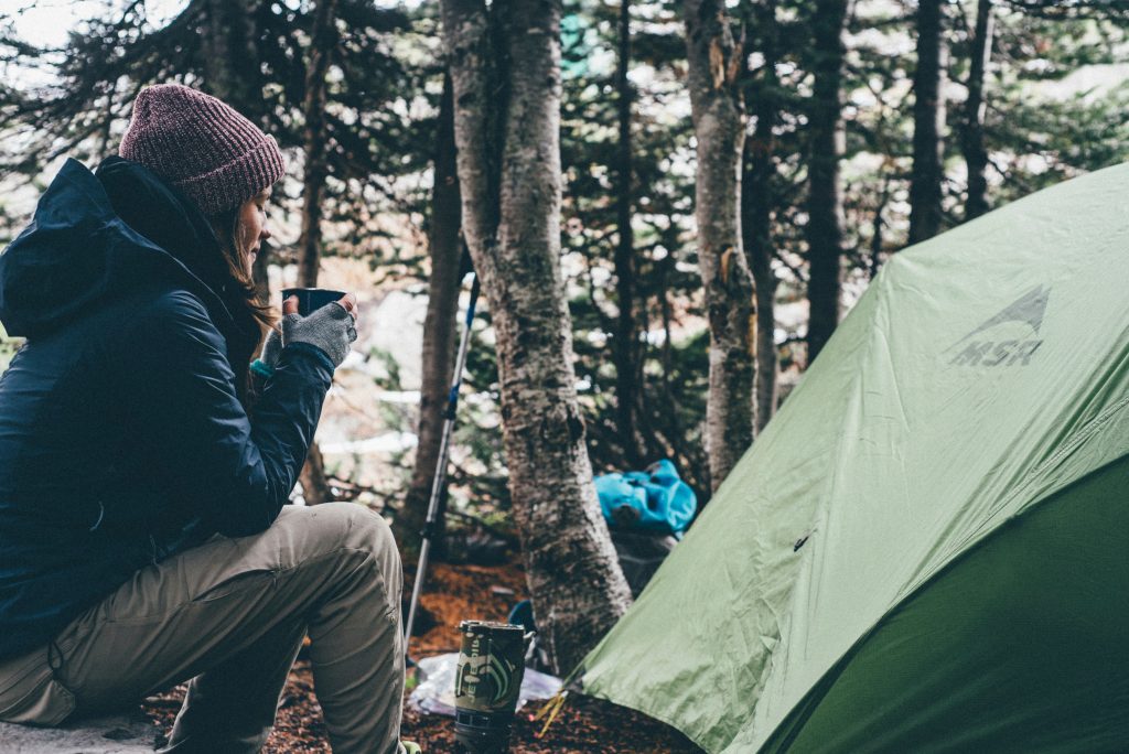 Woman in winter gear drinking a hot drink sat next to tent