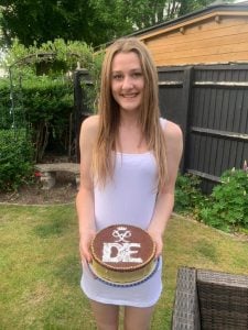 Young woman in white dress holding cake with DofE logo