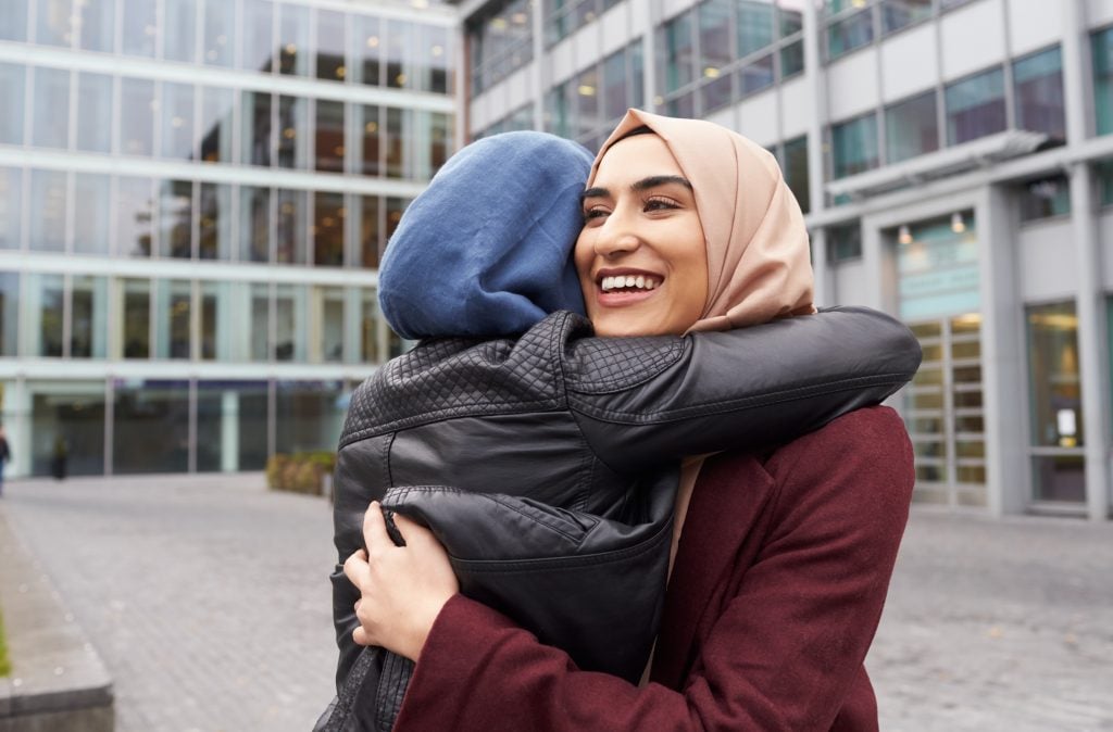 Two women in head scarves hugging in front of building and smiling