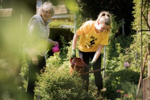 A young woman wearing a yellow "Do It 4 Youth" t-shirt and green gardening gloves is watering plants with a red watering can in a lush garden. Beside her, an older woman with short gray hair, wearing a patterned vest and dark trousers, smiles while holding a small pink watering can. The scene is bathed in sunlight, surrounded by greenery and blooming flowers, suggesting a community or intergenerational gardening activity.