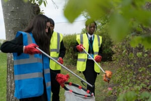 Three young people in school uniforms and high-visibility vests are participating in a litter-picking activity outdoors. They are wearing red gloves and using litter pickers to collect rubbish, placing it into a waste bag. One person in a blue vest is in the foreground, reaching down with a litter picker, while the other two, in yellow vests, are in the background, also picking up litter. The setting is a grassy area with trees and bushes.