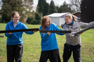 Three young girls are participating in an outdoor team challenge, holding sections of black guttering to transport water from one end to the other. They are dressed in warm clothing, with two wearing blue fleeces and one in a grey jacket. Water is visibly leaking from the gaps, and they are laughing and smiling as they work together. The background features a grassy field with tents and trees, suggesting a camp or outdoor activity setting.