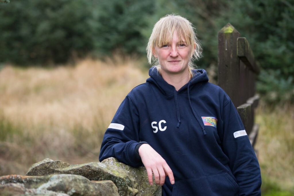 A D of E Leader with blonde hair and a navy hoodie leans on a stone wall.
