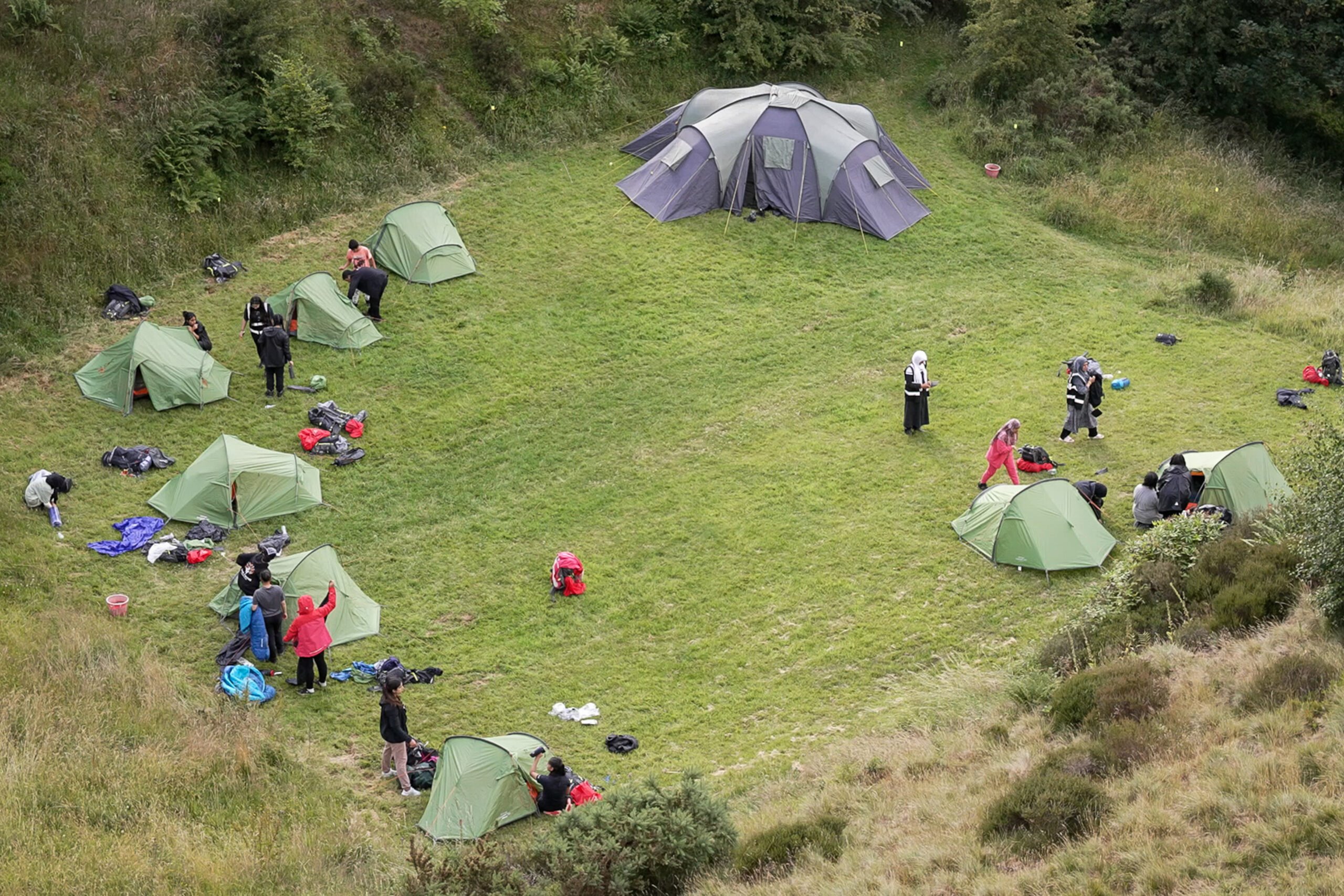 An aerial shot of a campsite. The tents are on grass, in a circle with a large space in the middle. There are young people next to their tents setting them up.