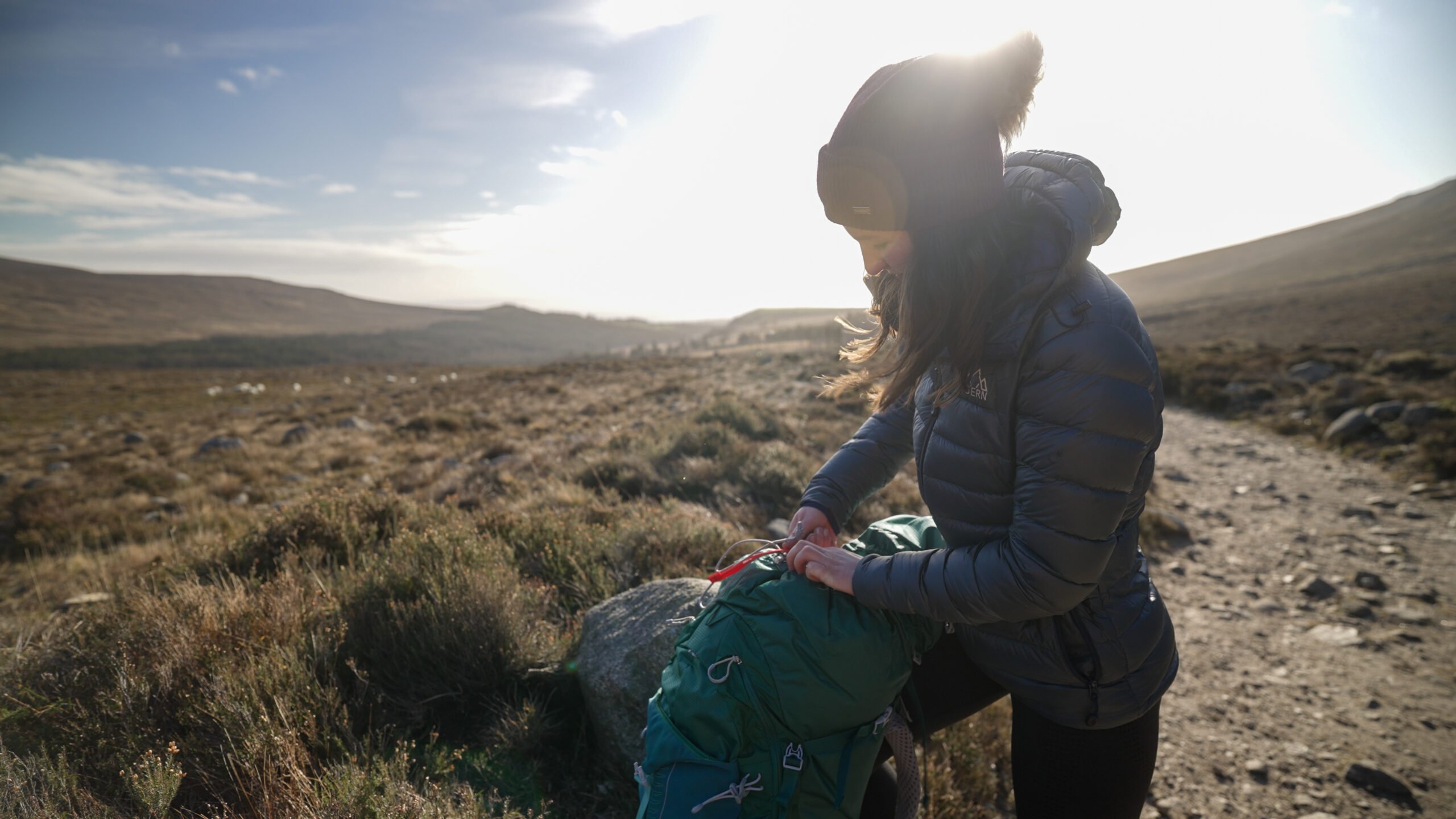 Young woman in a warm jacket and beanie adjusting her backpack on a countryside hiking trail, with open moorland and hills in the background at sunrise.