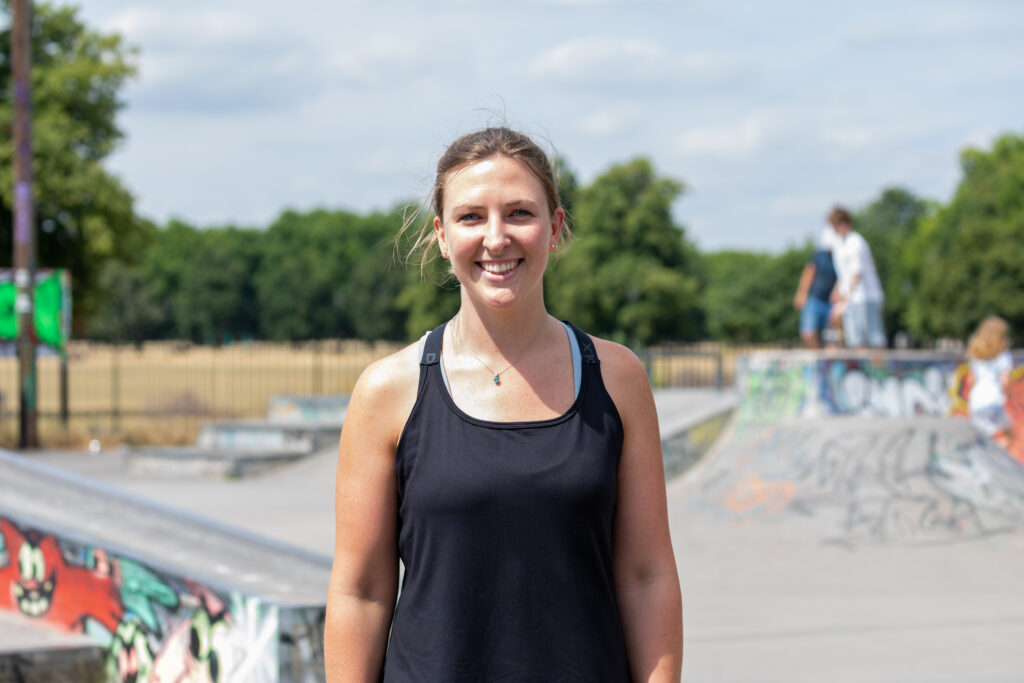 Jemma has brown hair and a black tank top, she's standing at a skatepark in the sun and smiling.