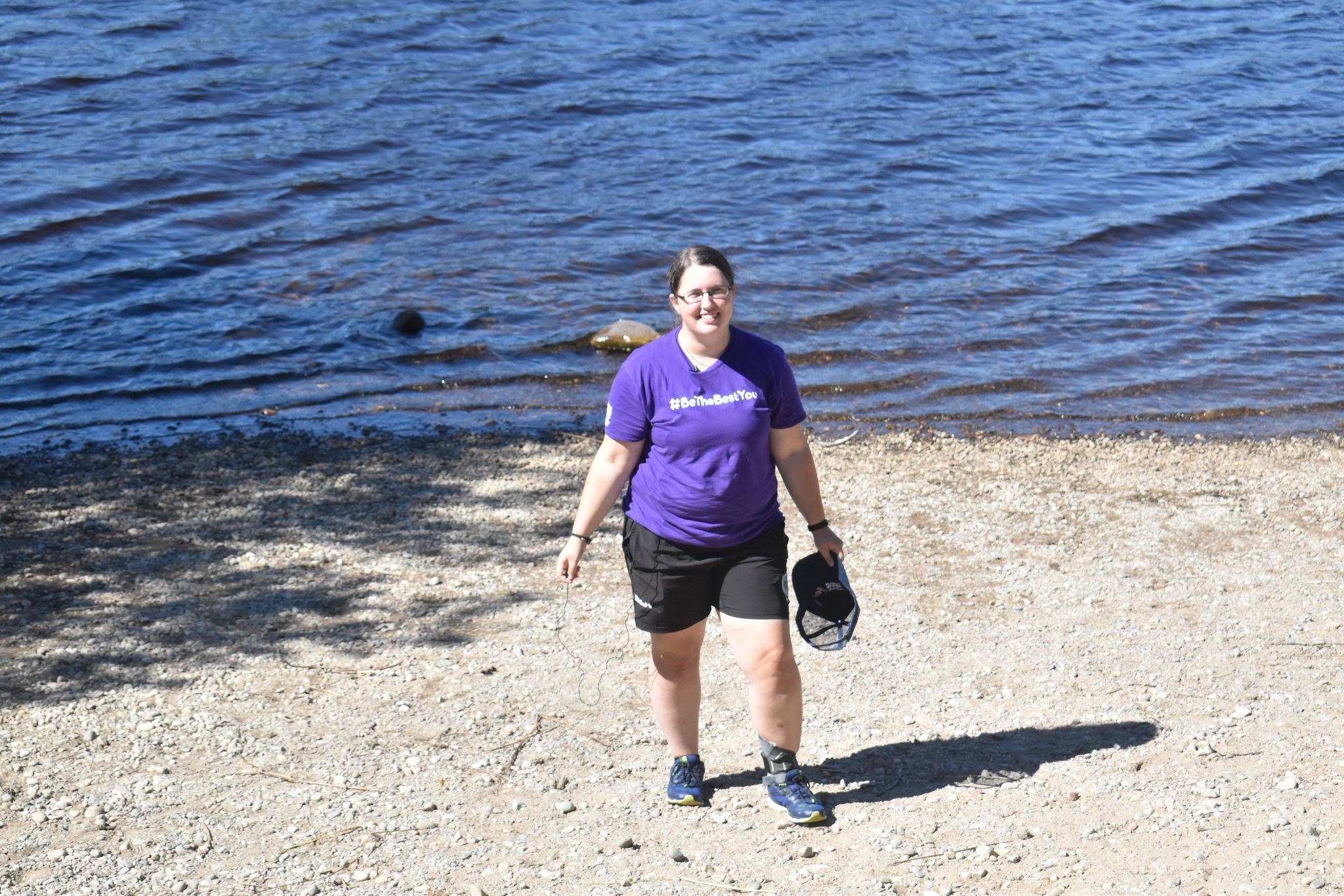 Anja, standing smiling in front of a body of water wearing a purple tshirt, black shorts, and holding a baseball cap