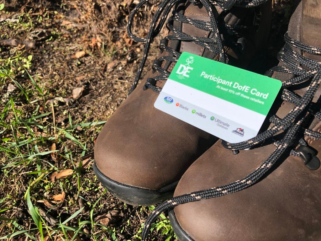 Pair of brown walking boots with green, resting on top of them is a white and green Participant DofE card which reads "At least 10% off these retailers: Go Outdoors, Blacks, Millets, Ultimate Outdoors and Tiso."
