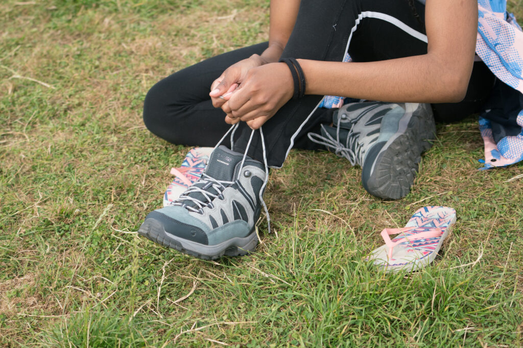 Hands tying the laces of grey walking boots while sitting on the grass with a pair of pink flipflops next to them.