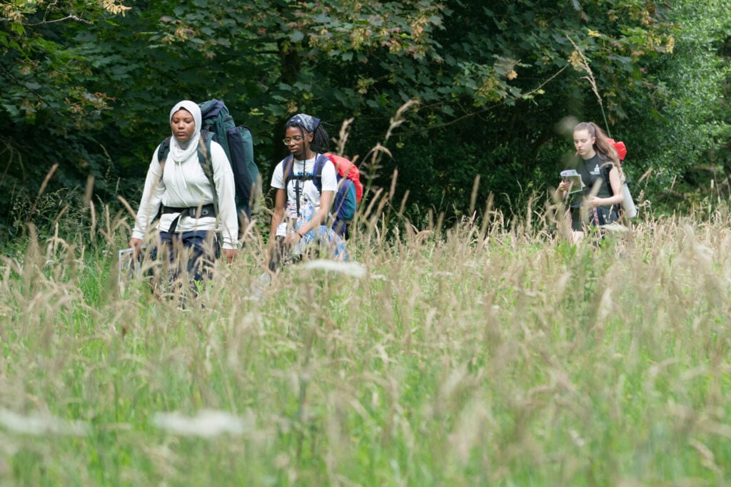 3 teenage girls wearing rucksacks, walking through long green grass reading a map during their DofE expedition.