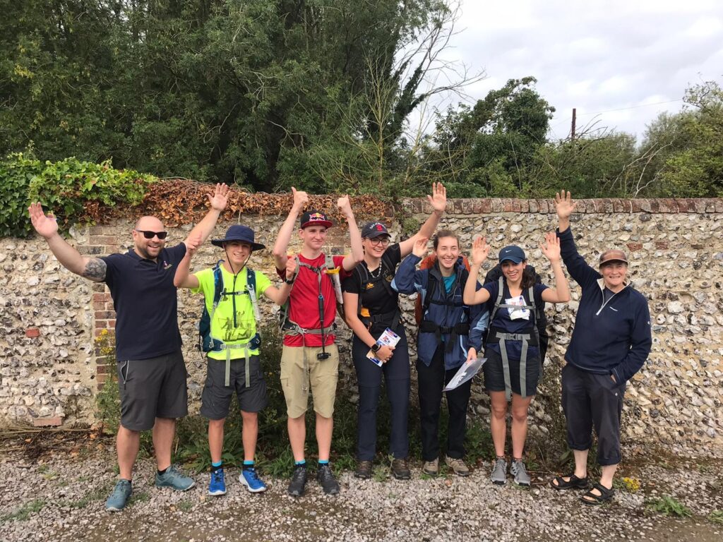 A group of seven young hikers stands in front of a stone wall, all smiling and raising their hands in the air. They are dressed in outdoor gear, including backpacks and hats, and appear to be celebrating or posing for a photo. The background features greenery and trees, suggesting a natural setting.