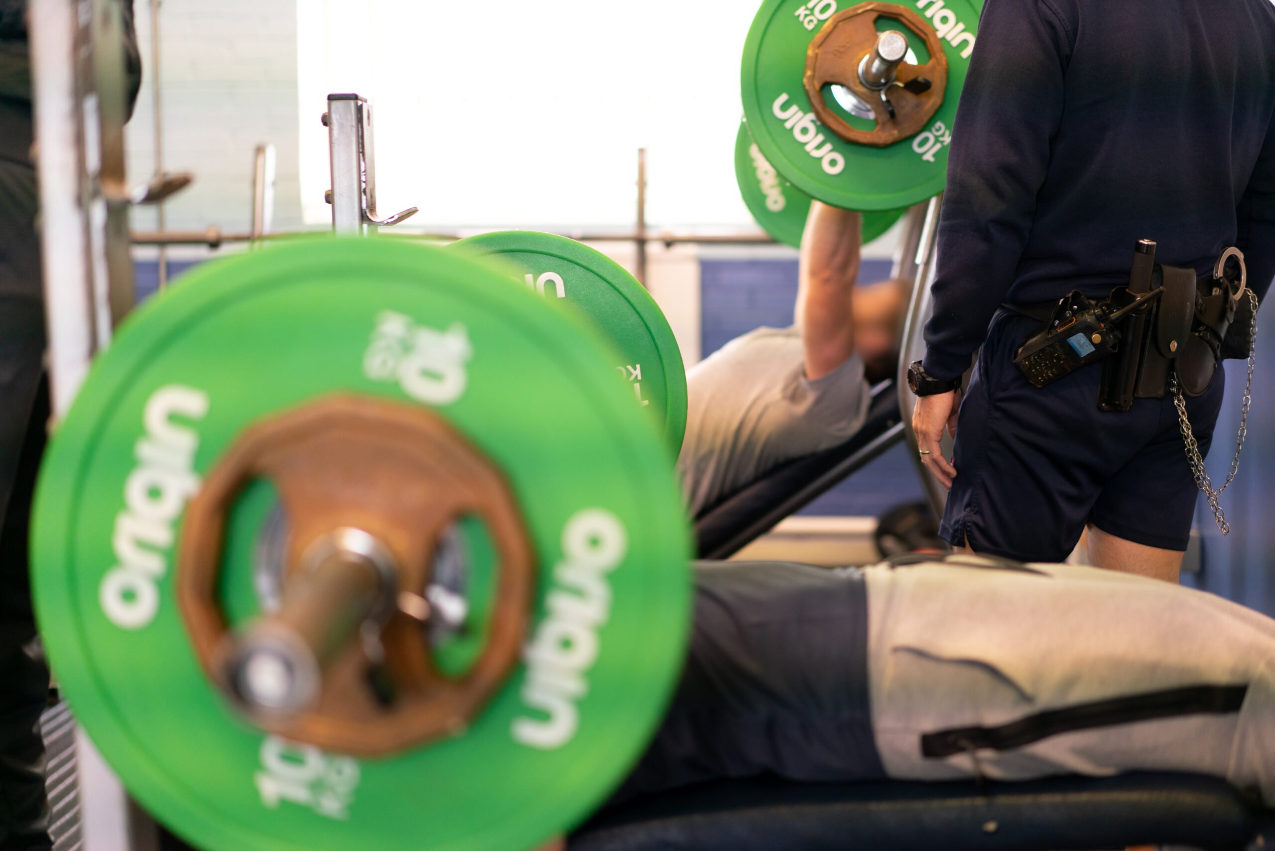 A young person lifting weights in prison. His face is obscured by the weight and we can see the prison officer in the background.