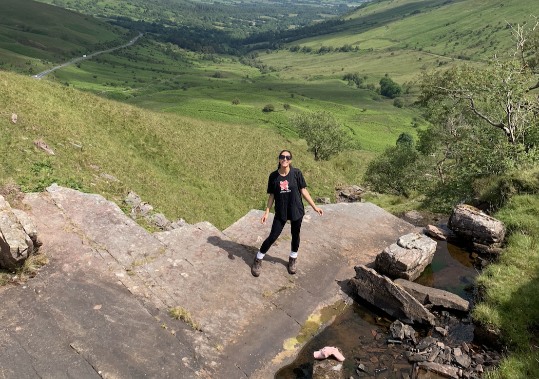 2023-24 UK Youth Ambassador Lauren. She is standing on the edge of a rock smiling to camera, in the background we can see green grass and hills.