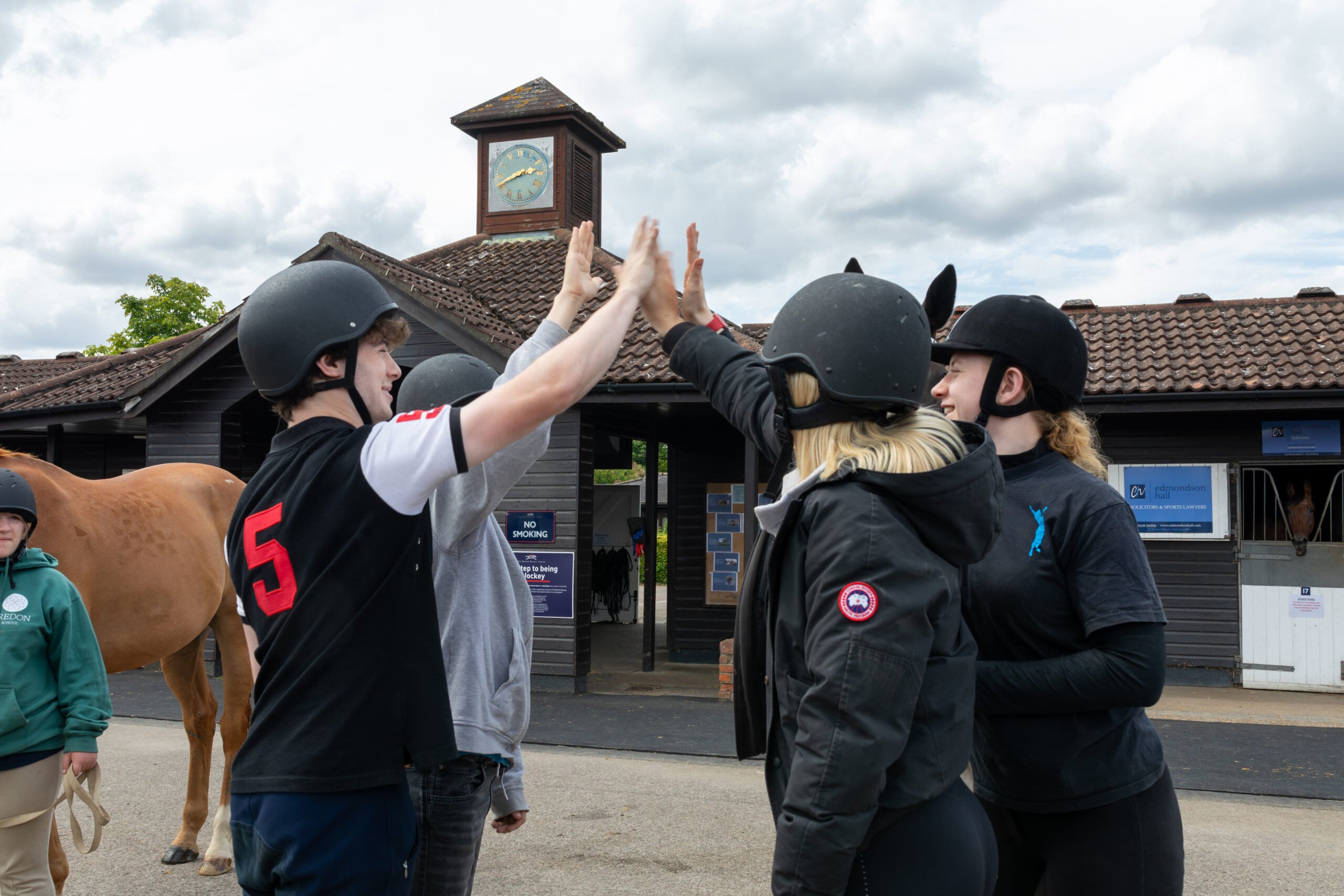 Alt text: A group of young people wearing helmets are giving high-fives outside a stable. In the background, there is a clock tower and a horse. The sky is partly cloudy.