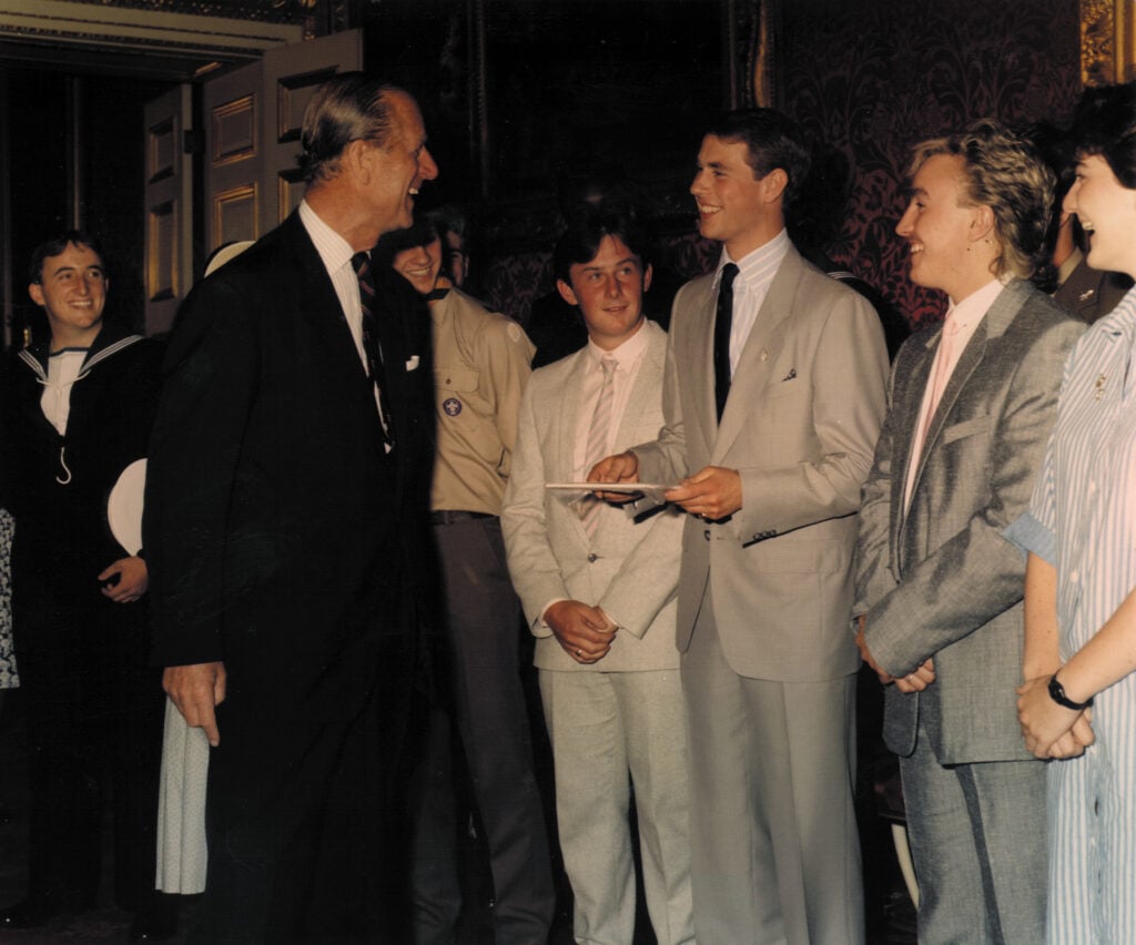 A group of people are gathered in a richly decorated room. An older gentleman in a dark suit is smiling and interacting with a younger man in a light grey suit who is holding an award. The young man appears to be in a formal ceremony. To the side, other young men and women, including one dressed in a naval uniform, are also present and looking on. The overall atmosphere seems formal and celebratory.