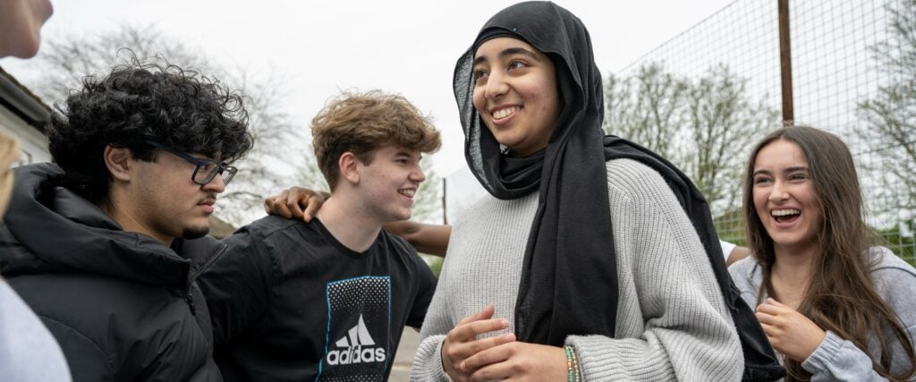 4 young people laughing together. They are standing outside and we can see trees and a high fence in the background.