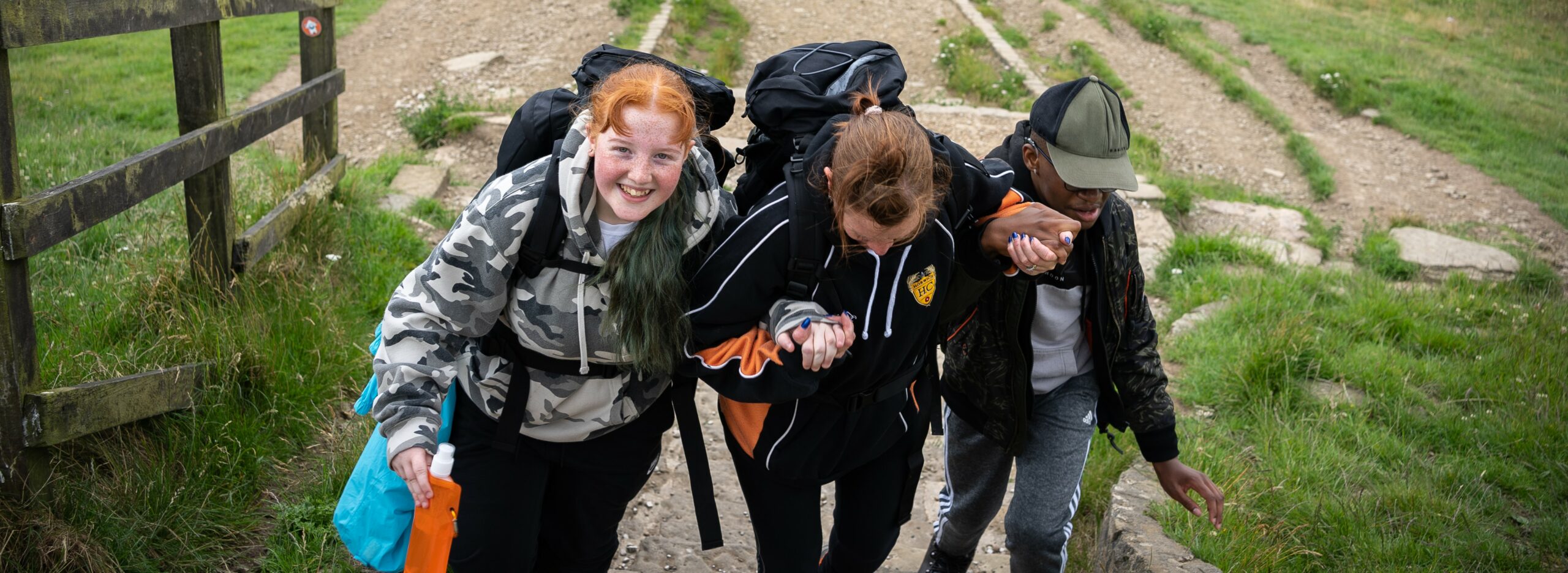 3 young people, all carrying camping equipment, hold hands as they walk up a hill. The person on the left is smiling to the camera and the other two people are looking down at their feet.
