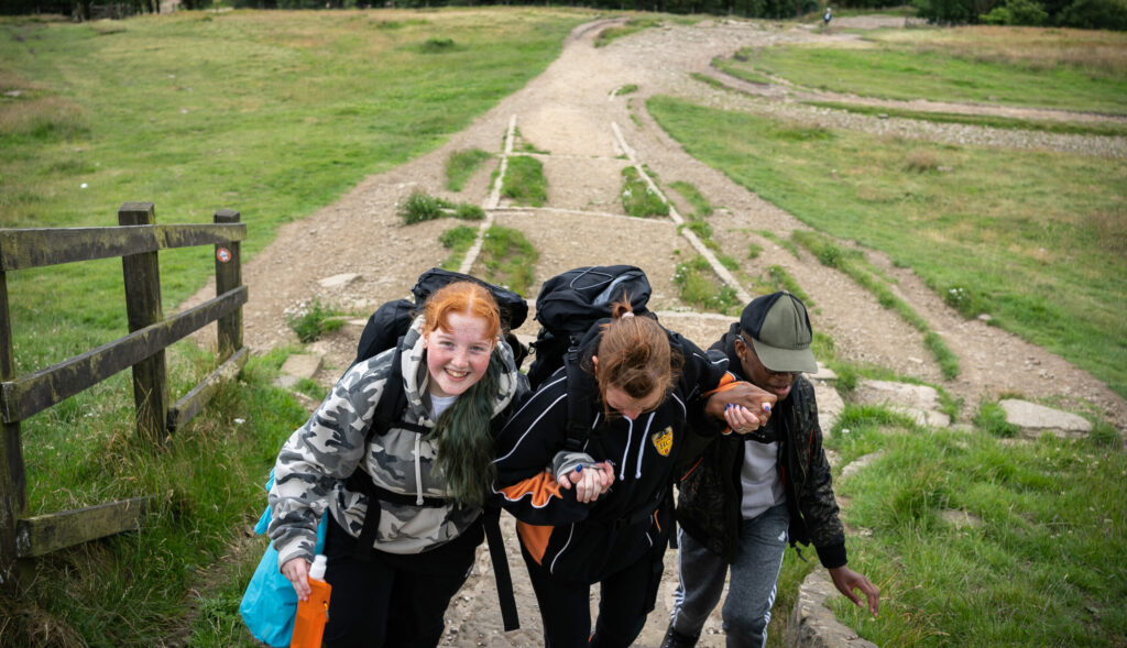 Three young people carrying rucksacks hold hands as they walk up a hill.