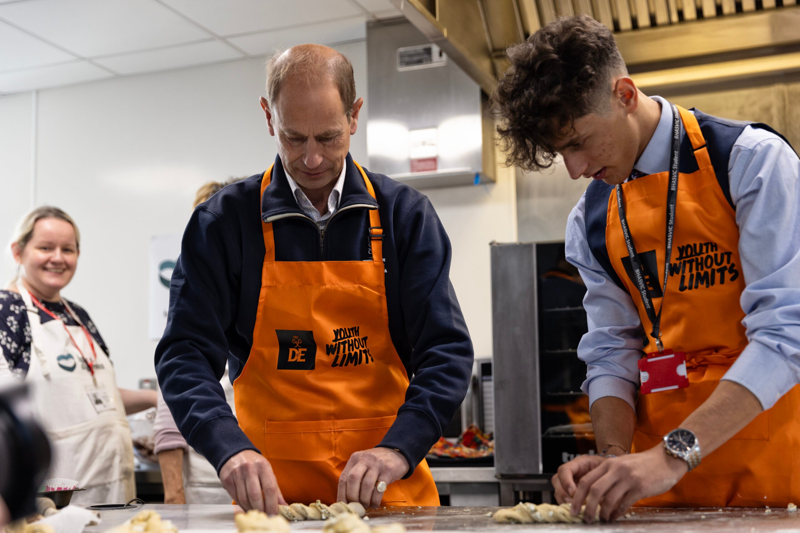 2 people kneading dough in an industrial kitchen. They are both wearing an orange apron that reads 'Youth Without Limits' and in the background a woman is smiling while watching them.