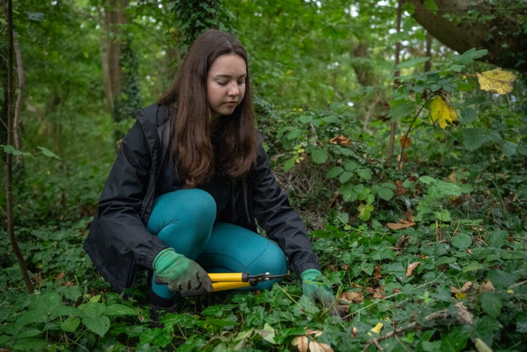 Girl with brown hair kneeling outside, in a forest setting, using loppers