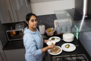 Sharandeep stands in a modern kitchen preparing a meal. She holds a small white plate with a piece of butter in one hand and a spoon in the other, about to spread the butter on a plate of food. The kitchen counter has several plates with traditional Indian food, including green saag (a spinach-based dish), butter, yogurt, and stacked flatbreads (rotis). Behind her, there is a microwave, a basket of fruit, and labeled storage jars for tea, sugar, and coffee. She looks towards the camera with a neutral expression.