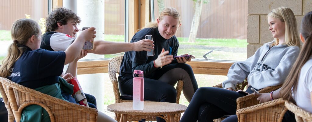 A group of young people sitting around a table showing each other their phone screens and smiling.