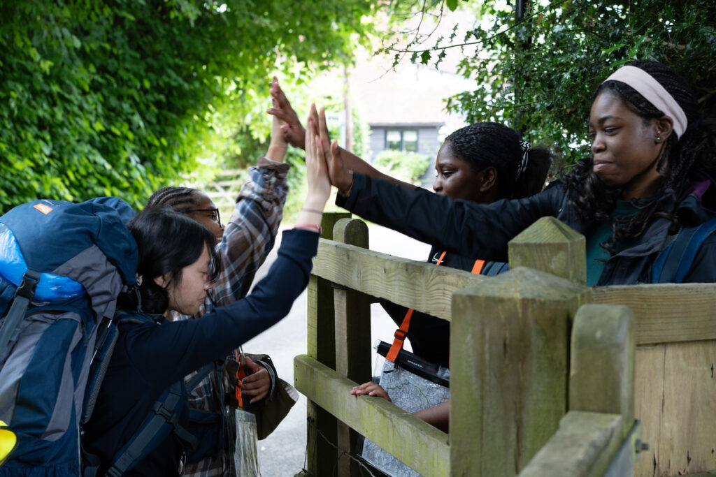 Young people on an expedition carrying rucksacks high five each other over a wooden gate.
