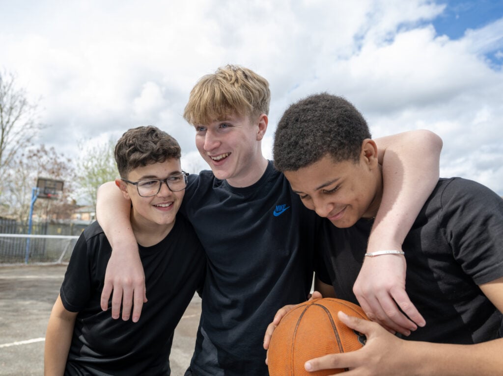 Three young people smiling outside, the middle one has their arms around the other two, one is holding a basketball.