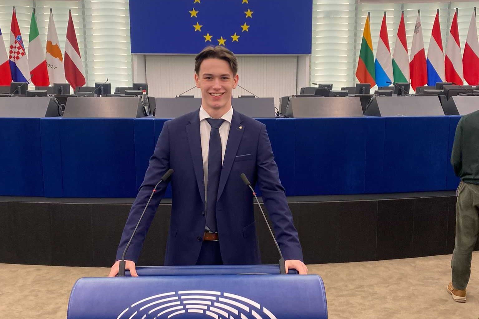 Jake in a dark blue suit, white shirt, and tie stands confidently at a podium in a large conference room. The podium features the emblem of the European Parliament. Behind him are rows of empty desks, microphones, and a blue flag with yellow stars symbolizing the European Union. Several national flags are visible in the background." "A young man in a dark blue suit, white shirt, and tie stands confidently at a podium in a large conference room. The podium features the emblem of the European Parliament. Behind him are rows of empty desks, microphones, and a blue flag with yellow stars symbolizing the European Union. Several national flags are visible in the background. 