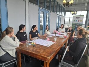 UK Youth Ambassaqdors are gathered around a rustic wooden table in a well-lit indoor space with large windows and blue-framed doors. One woman, wearing a black shirt and beige pants, is speaking and gesturing with her hand while others listen attentively. The table is covered with papers, sticky notes, pens, notebooks, water bottles, and small pink candles. A banner in the background displays the hashtag '#WORLD READY' along with icons and an image of a person in a field. The participants are dressed in casual and semi-formal attire, engaged in discussion.