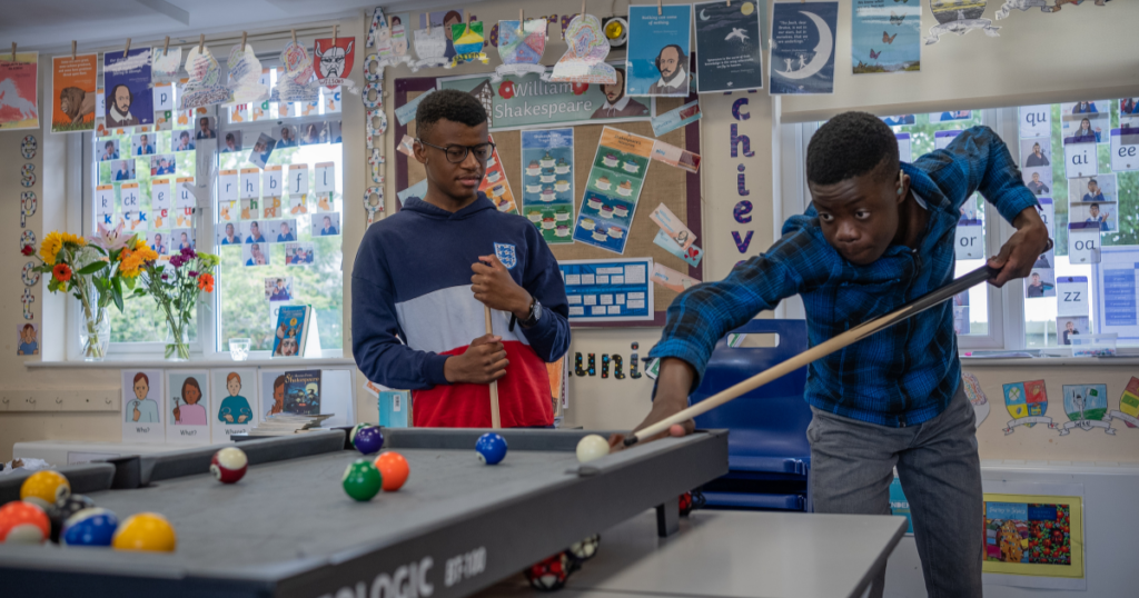 Gio and Prince play a game of pool in a classroom setting. One, wearing a blue plaid shirt, leans forward intensely, lining up a shot with his cue stick. The other, wearing a navy and red hoodie with an England emblem, stands nearby holding a cue stick, watching with a thoughtful expression. The classroom walls are decorated with educational posters, student artwork, and images related to William Shakespeare. A bulletin board displays various learning materials, and large windows let in natural light. A vase with colorful flowers sits on a table near the windows.