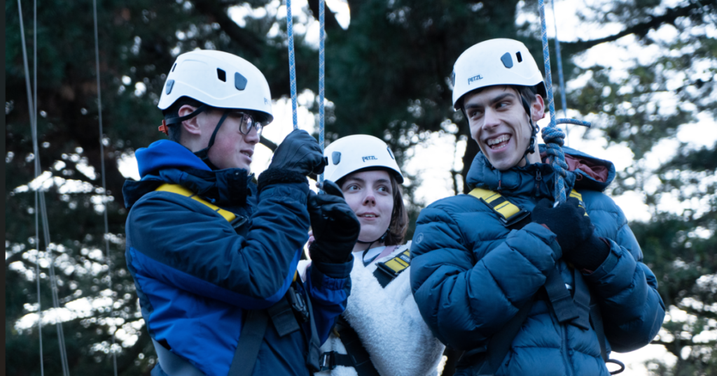 Three individuals wearing helmets and harnesses are standing together outdoors, preparing for an activity that involves ropes. They are smiling and engaged in conversation, with trees in the background. The scene suggests a sense of adventure and teamwork.