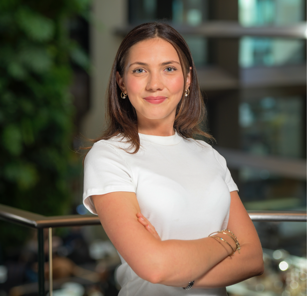 A young woman with shoulder-length brown hair and light skin stands with her arms crossed, smiling confidently. She is wearing a white short-sleeved top, gold hoop earrings, and gold bracelets. The background is blurred, featuring a modern office setting with glass railings, green plants, and people working in the distance.