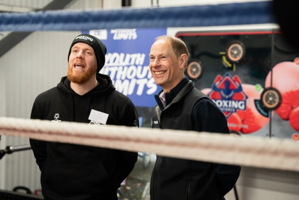 Joe Osman, wearing a black hoodie with a logo and a black beanie, is speaking while standing next to HRH The Duke of Edinburgh who is smiling. The Duke is dressed in a black vest over a dark sweater. They are inside a boxing gym, with a boxing ring rope visible in the foreground. The background features a blue banner with the words "YOUTH WITHOUT LIMITS" and a boxing-themed interactive training wall with red gloves and target pads.