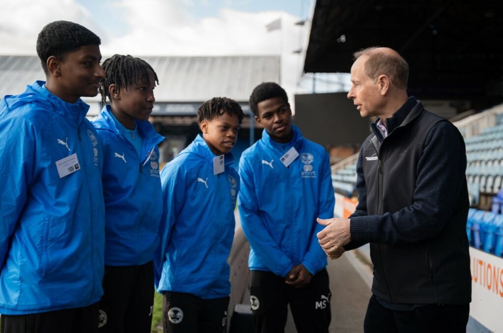 HRH The Duke of Edinburgh is engaging in conversation with four young people wearing matching blue Puma-branded jackets with the "SYTFC" logo. The young people, all wearing name tags, stand attentively, listening and smiling. The setting is an outdoor football stadium with seating and a partially covered stand visible in the background. The Duke is wearing a black fleece vest over a dark sweater and gestures with his hands as he speaks. The interaction appears to be part of a formal visit or event.