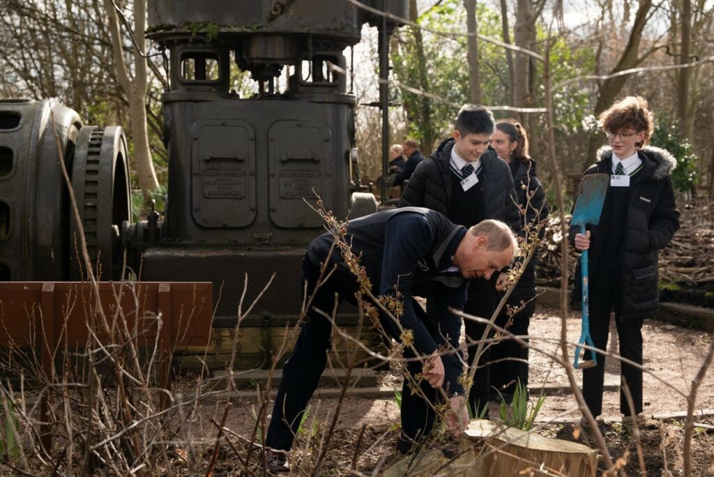 HRH The Duke of Edinburgh is bending down to plant a small tree or shrub in an outdoor setting, surrounded by young DofE participants. The young people, dressed in school uniforms with black coats and name tags, watch attentively, with one holding a spade. The background features an old industrial machine, leafless trees, and a natural woodland environment, suggesting a conservation or environmental activity at Railworld Wildlife Haven. The scene is lit by natural sunlight.