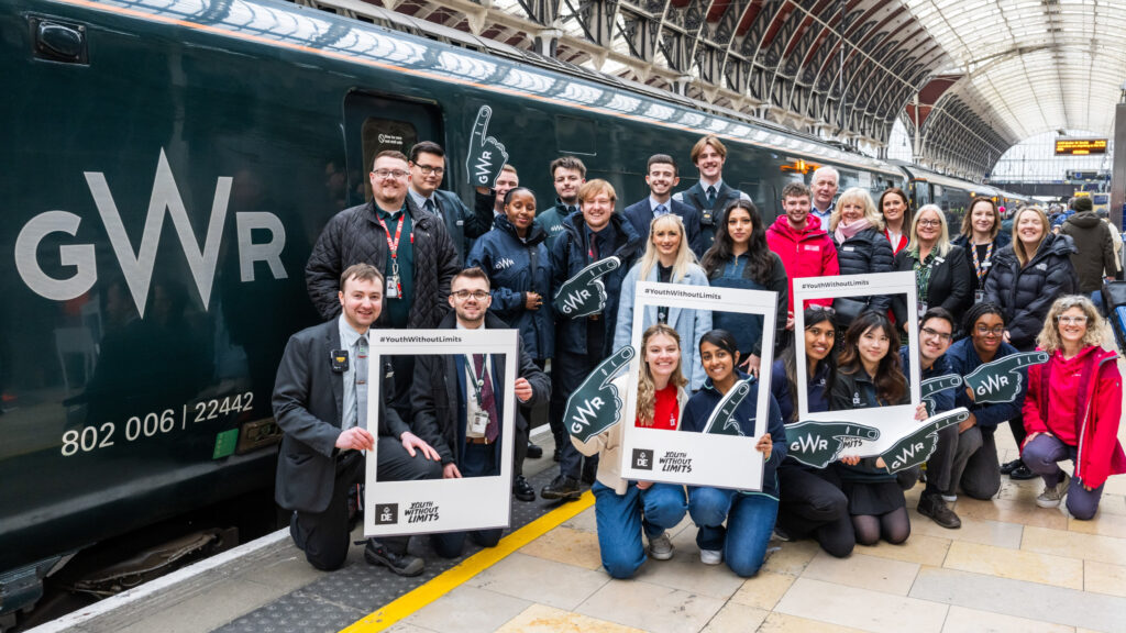 A large group of people, including young apprentices and railway staff, pose together on a train platform in front of a Great Western Railway (GWR) train. Many are holding signs and photo frames with the hashtag '#YouthWithoutLimits' and the GWR logo. The group is diverse, with individuals wearing uniforms, business attire, and casual clothing. The setting is a well-lit railway station with a high arched ceiling and a digital departure board visible in the background.