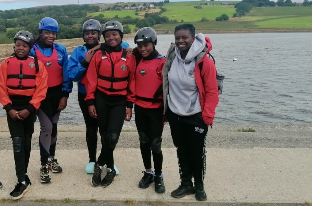 A group of six young people stands by a body of water, wearing helmets and life jackets. They are dressed in athletic clothing suitable for water activities. The background features green hills and a cloudy sky, indicating an outdoor setting. The group appears to be smiling and enjoying their time together.