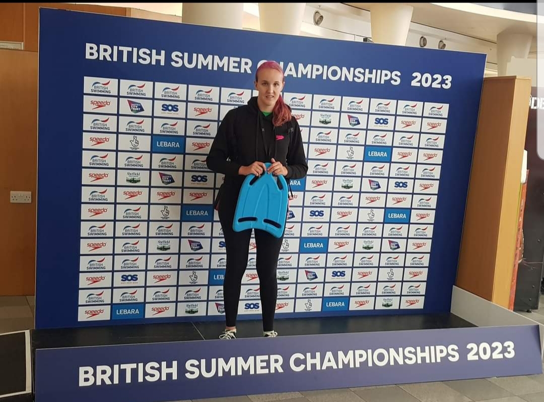 Abi stands on a podium at the British Summer Championships 2023. She is wearing a black tracksuit and holding a blue kickboard. Behind her is a backdrop featuring the event name and sponsor logos, including British Swimming and Speedo. She has a proud and confident expression as she looks at the camera.