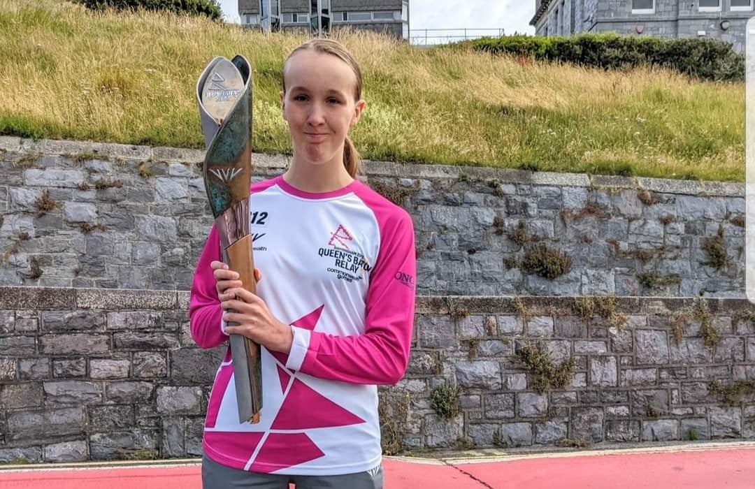 Abi stands on a running track, holding the Queen’s Baton from the Queen’s Baton Relay. She wears a pink and white relay shirt with the event logo and the number 12 printed on it. She has a proud and happy expression as she looks at the camera. Behind her is a stone wall with grass and buildings visible at the top.