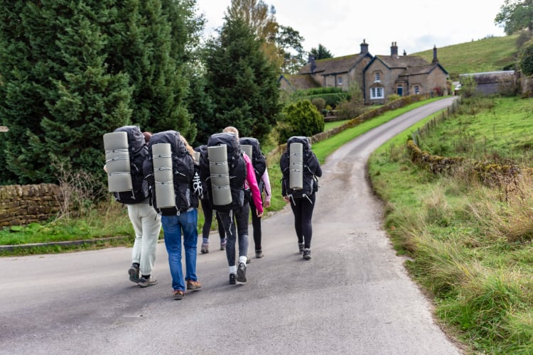 Six hikers, each carrying a large backpack, walks along a winding country road. They are heading towards a stone house on a hillside, surrounded by greenery and trees. The sky is partly cloudy, and the landscape is lush and inviting.