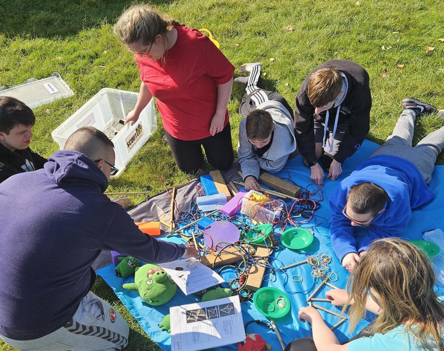 A group of six people is gathered on a blue tarp in a grassy outdoor setting. They are engaged in an activity involving various colorful materials, including wires, papers, and small green objects. Some individuals are sitting while others are kneeling, all focused on the task at hand. The scene is bright and sunny, with trees and a few people visible in the background.