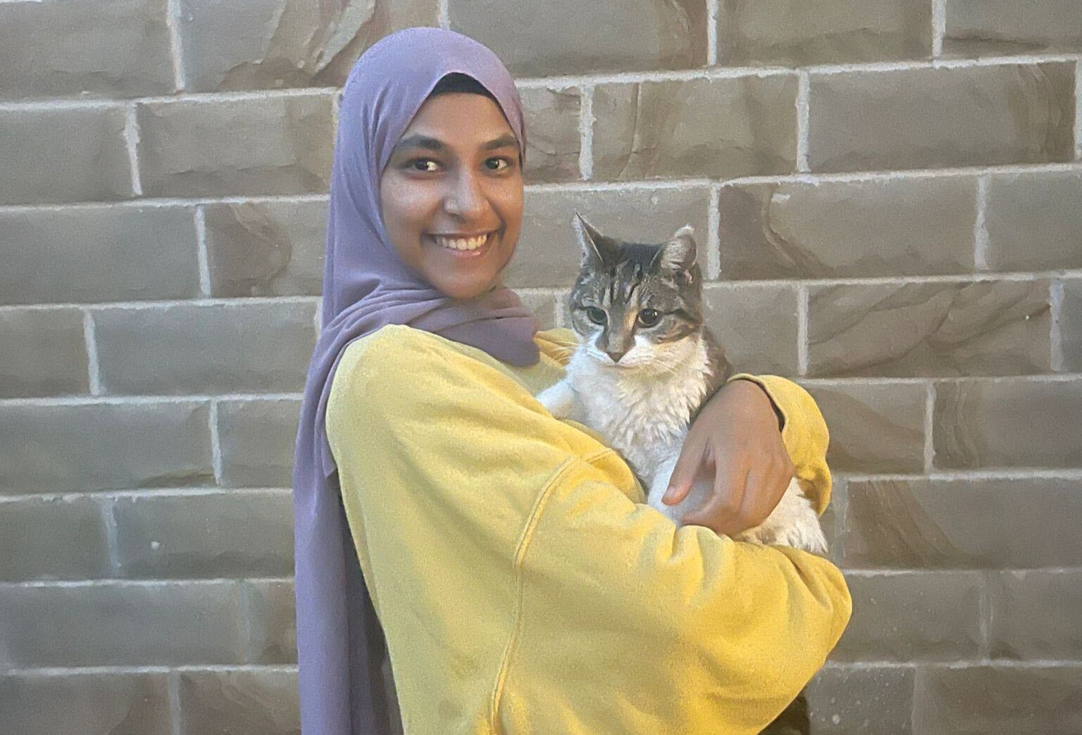 Safa, wearing a lavender hijab and a yellow oversized sweatshirt is smiling while holding a fluffy cat in her arms. The cat has a mix of white and grey fur with darker tabby markings on its head and ears. It looks slightly serious as it gazes into the distance. The background consists of a neutral-toned brick wall. Safa's expression conveys warmth and happiness as she gently cradles the cat.