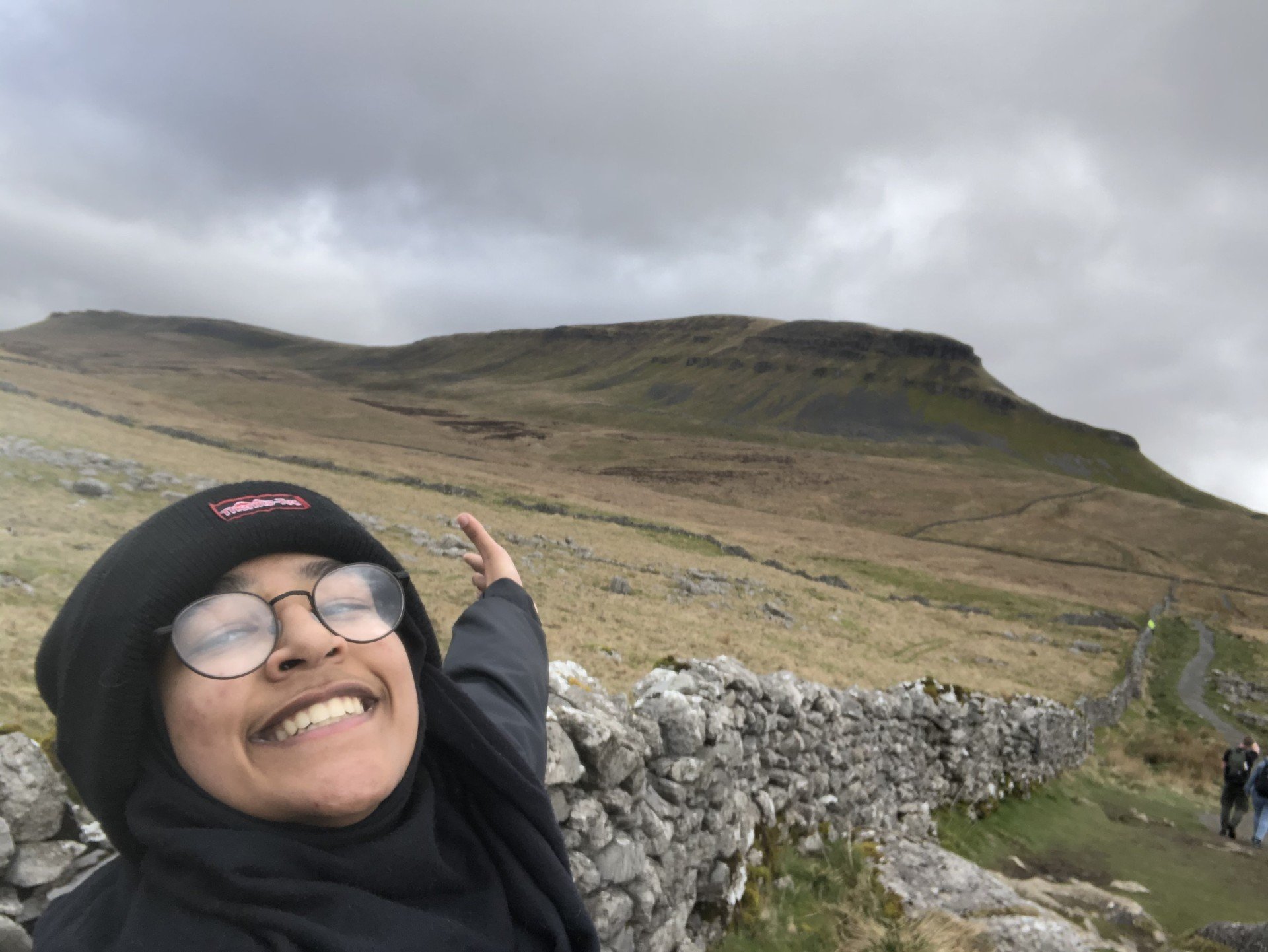 Safa is taking a selfie while hiking in the countryside. She is wearing a black beanie with a red logo, round glasses, and a black scarf. She has a big smile on her face and is extending one arm as if presenting the scenic landscape behind her. The background features a rugged hill with a flat top, covered in patches of green and brown vegetation, under an overcast sky. A winding stone wall lines the trail, and a few other hikers can be seen in the distance.