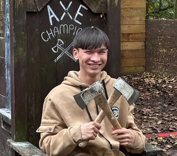 Evan holding two axes in a cross, sitting in a wooden throne that says "Axe Champion".