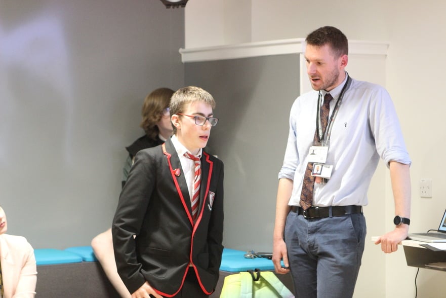 A young person in a school uniform presenting, standing next to a teacher leaning on a table.